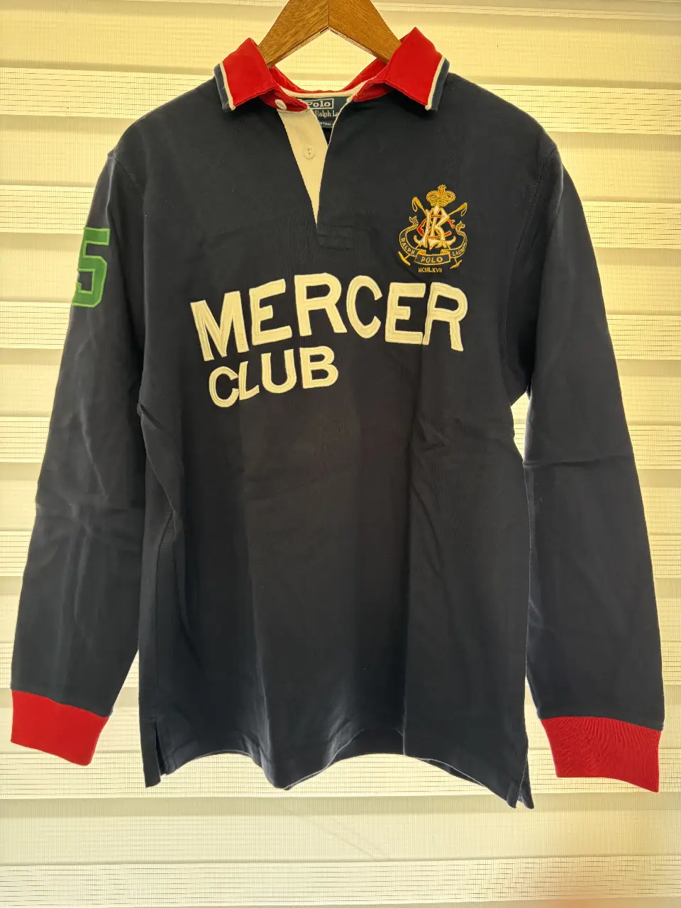 Remera manga larga de hombre Polo by Ralph Lauren, modelo Mercer Club. Confeccionada en algodón, de color azul marino con detalles en rojo y blanco. Presenta el logo "Mercer Club" en el frente y el número 5 en la manga izquierda. Cuello tipo polo con botones.