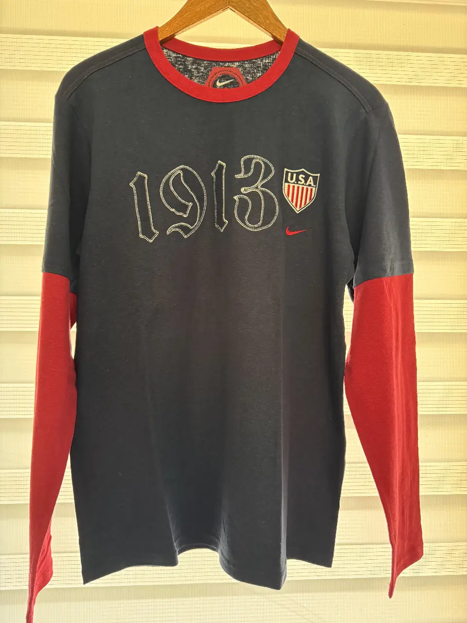 Remera manga larga de hombre, con mangas rojas y cuerpo azul marino. Estampado "1913" y logo de Nike en blanco. Diseño retro con detalles de costura. Ideal para un look casual y deportivo.