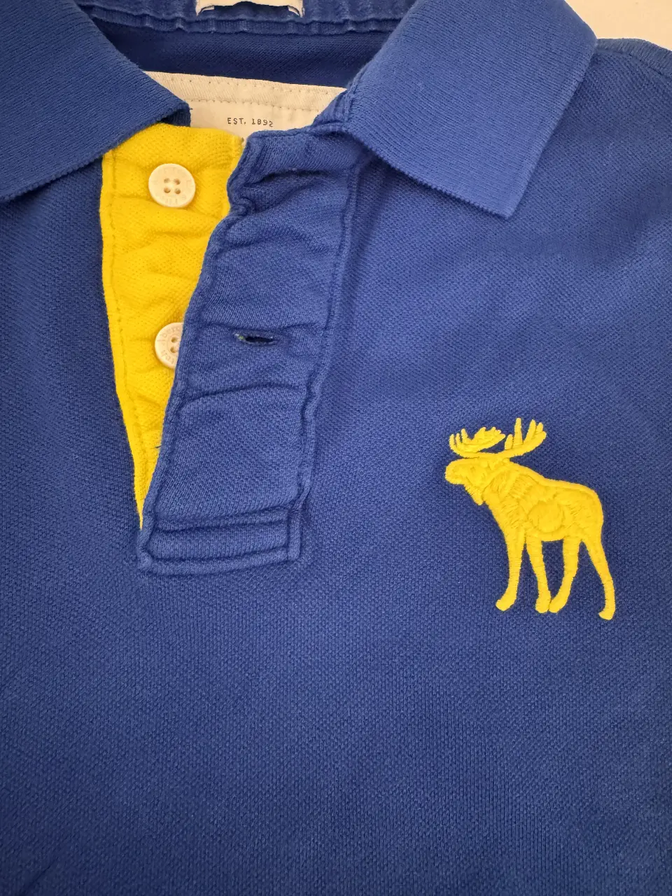 Remera polo azul de hombre, con detalles en amarillo en el cuello y logo de alce bordado. Marca Abercrombie & Fitch, talle M.