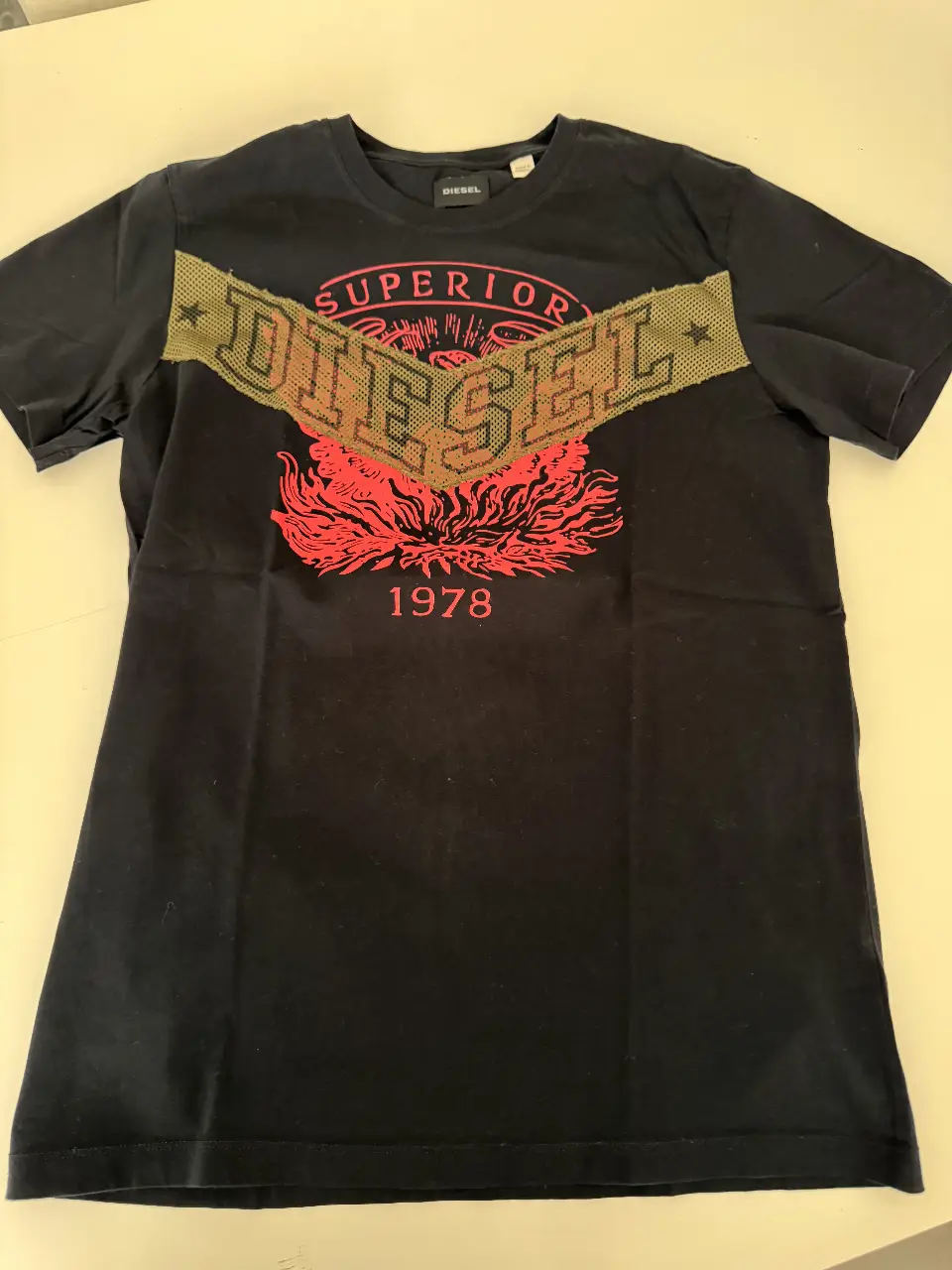 Remera Diesel negra con estampado frontal en rojo y dorado. Diseño con logo de la marca y detalles vintage. Ideal para un look casual y con estilo.