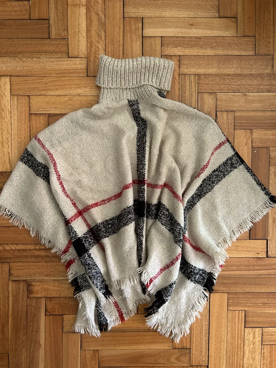Poncho de lana tejido a cuadros con cuello alto y flecos. Un clásico infalible para los días fríos, ideal para sumar un toque chic a cualquier look.