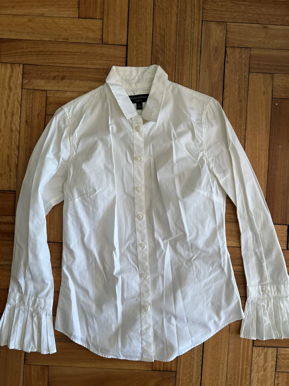 Camisa blanca de vestir con mangas largas y detalles plisados en los puños. Un clásico que no puede faltar en tu guardarropa, ideal para looks formales o para darle un toque chic a tu outfit diario.