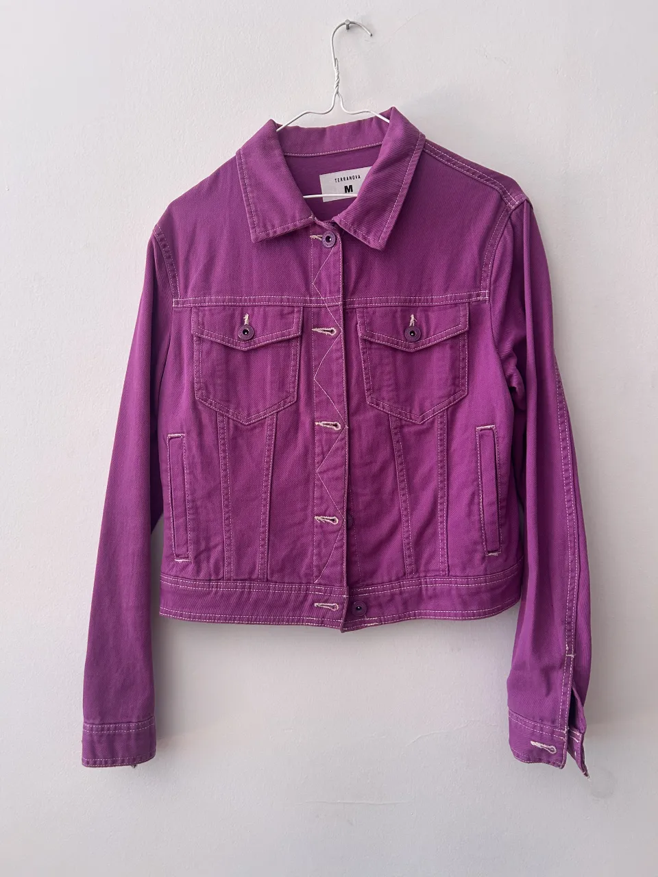 Campera de jean color violeta con pespuntes blancos. Tiene cuello solapa, botones al frente y en los puños, y bolsillos con tapa en el pecho.