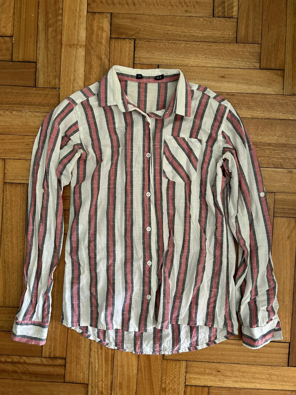 Camisa a rayas blanca, roja y gris. Manga larga con posibilidad de enrollar y sujetar con un botón. Cuello camisero y un bolsillo en el pecho.