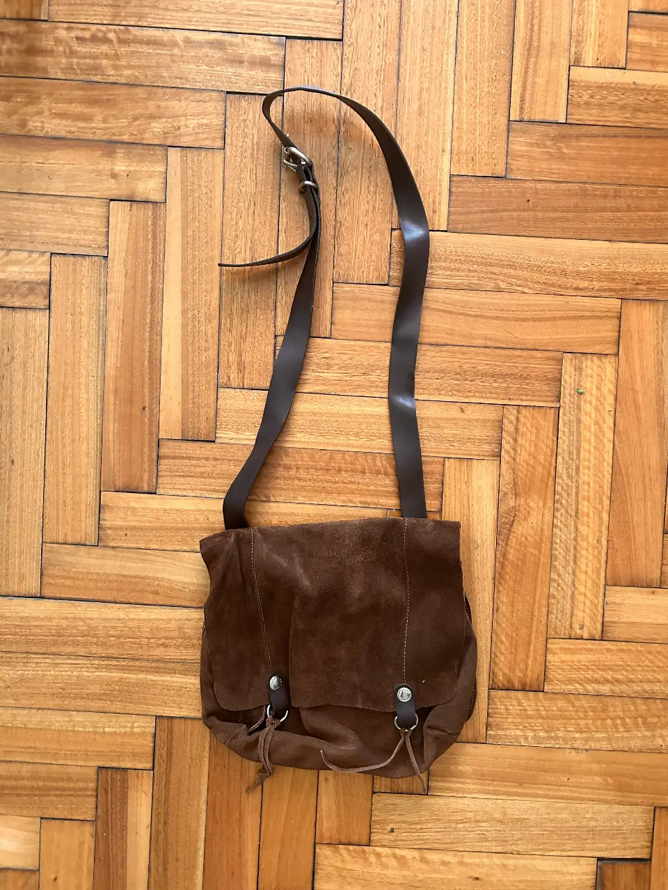 Bandolera de gamuza marrón, con solapa y cierre con tientos. Interior con bolsillo con cierre y etiqueta de la marca. Correa regulable.