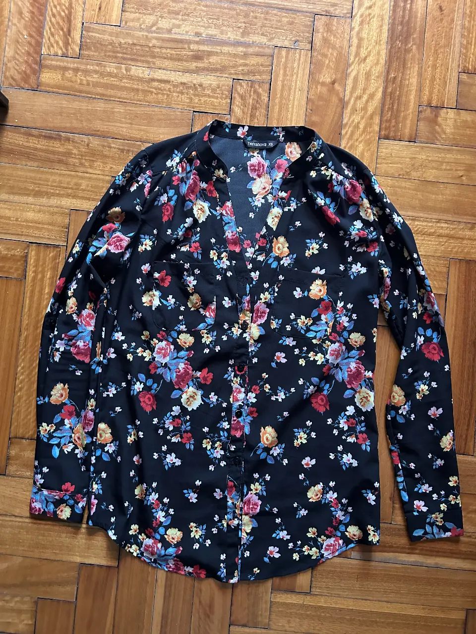 Camisa de mujer con estampado floral en tonos rojos, amarillos y azules sobre fondo negro. Cuello en V y mangas largas. Ideal para un look casual y femenino.