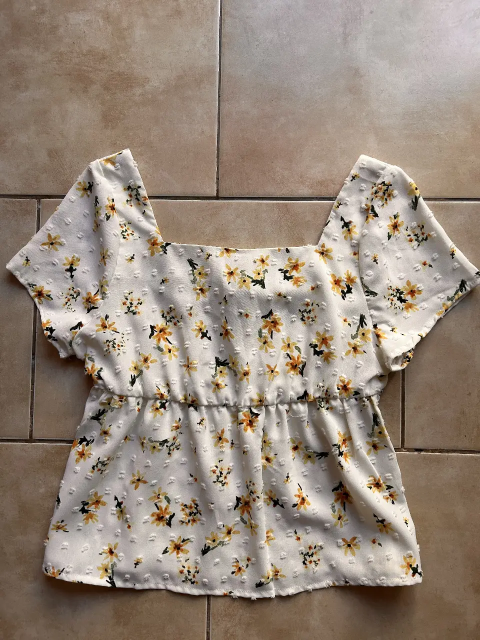 Blusa de mujer con estampado floral en tonos amarillos sobre fondo blanco. Diseño con corte cuadrado en el cuello y mangas cortas. Detalle de fruncido en la cintura. Ideal para un look fresco y primaveral.