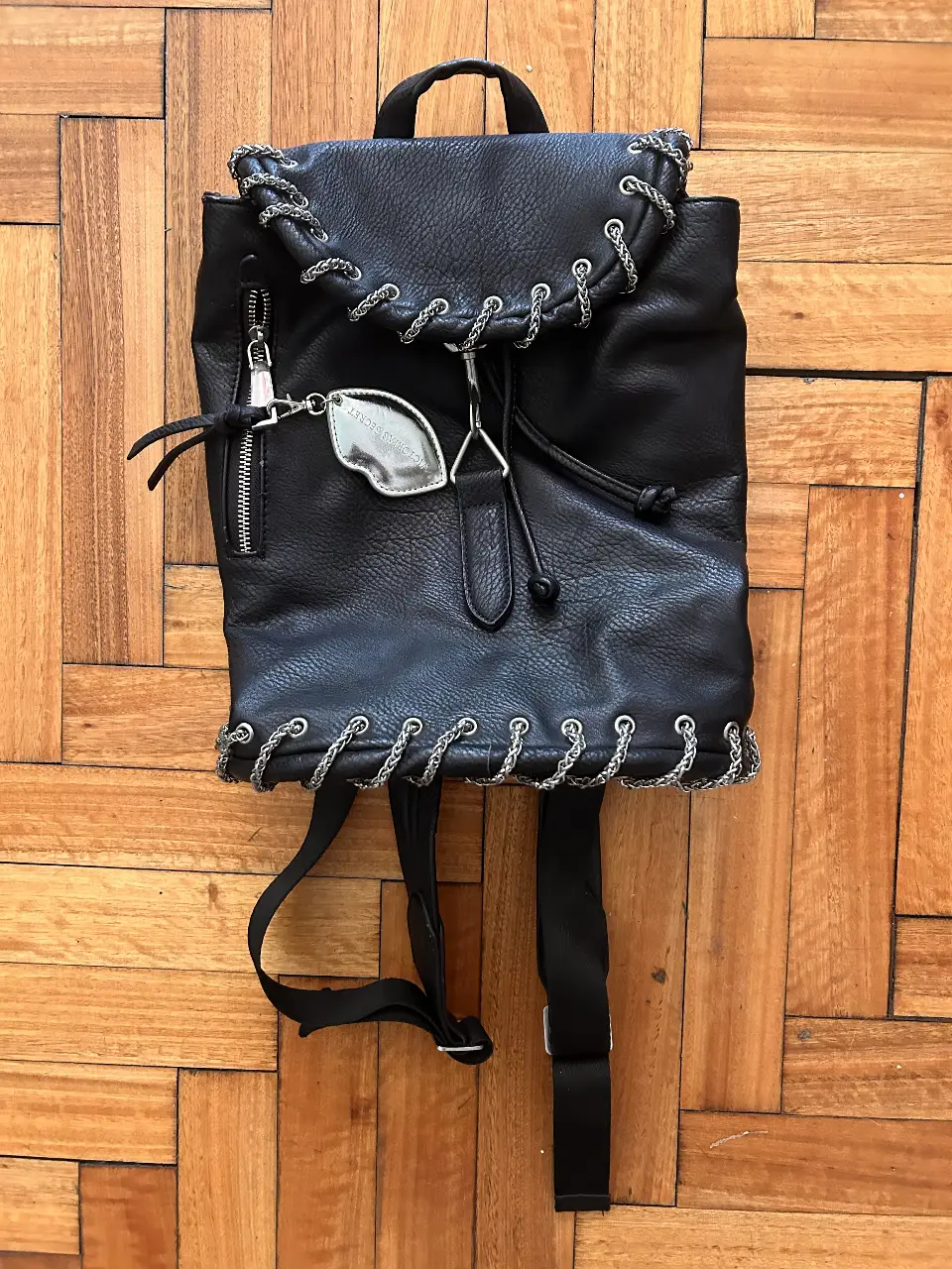 Mochila negra de cuero sintético con detalles de cadena plateada. Tiene un bolsillo frontal con cierre y un dije metálico. La solapa superior se cierra con un cordón. En el interior, tiene una etiqueta con la marca KOSIUKO. Ideal para un look urbano y moderno.