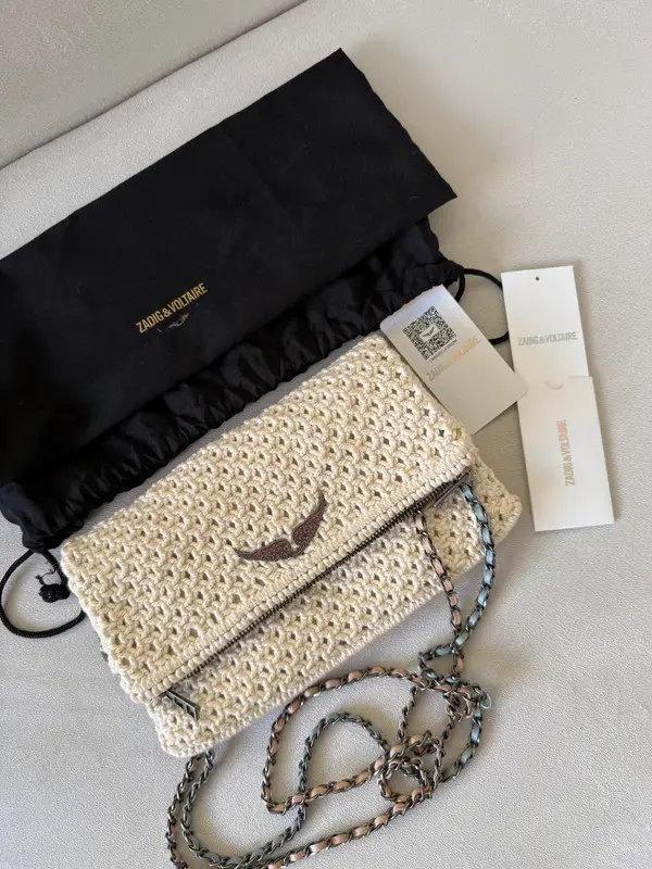 Cartera ZV bandolera de tejido artesanal en color crudo con detalles plateados. Diseño con solapa y cierre, cadena para colgar y etiqueta de la marca. Ideal para un look casual y con estilo.