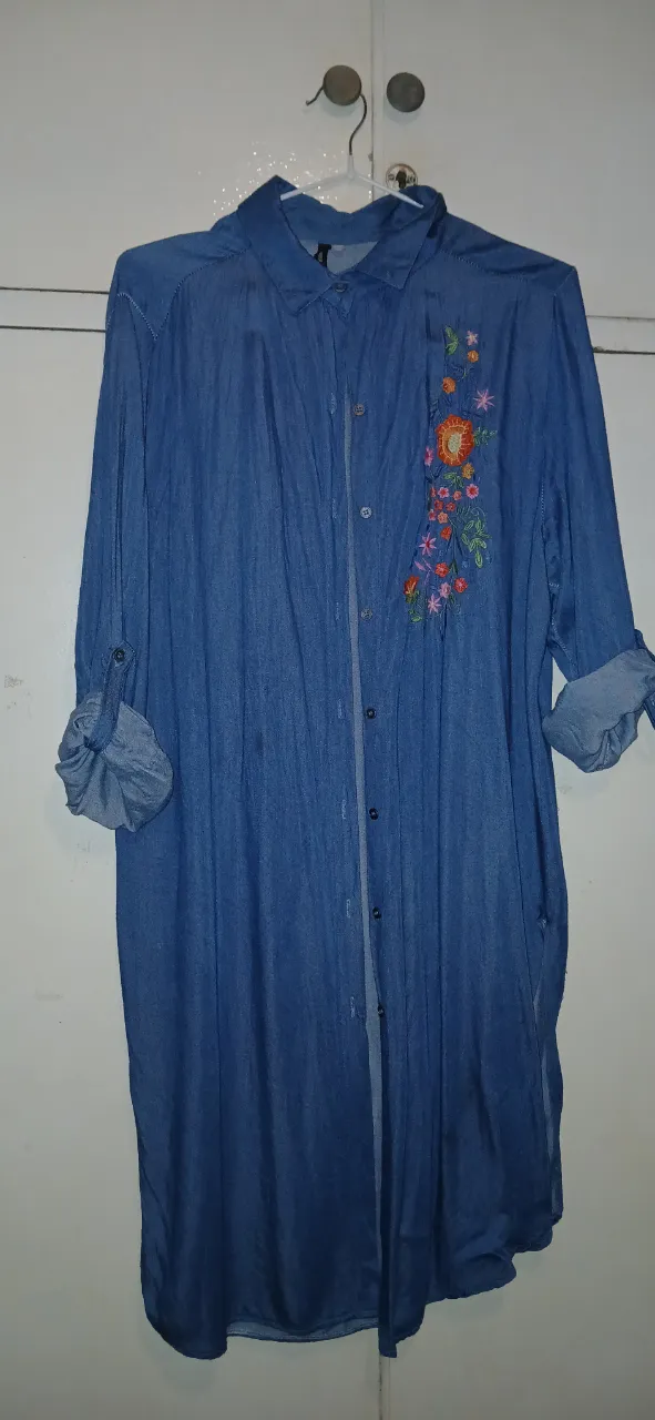 Camisa larga de jean con botones al frente y bordado floral en la zona del busto. Las mangas se pueden enrollar y ajustar con un botón. 40cm de hombros, 49cm de busto y 98cm de largo total.