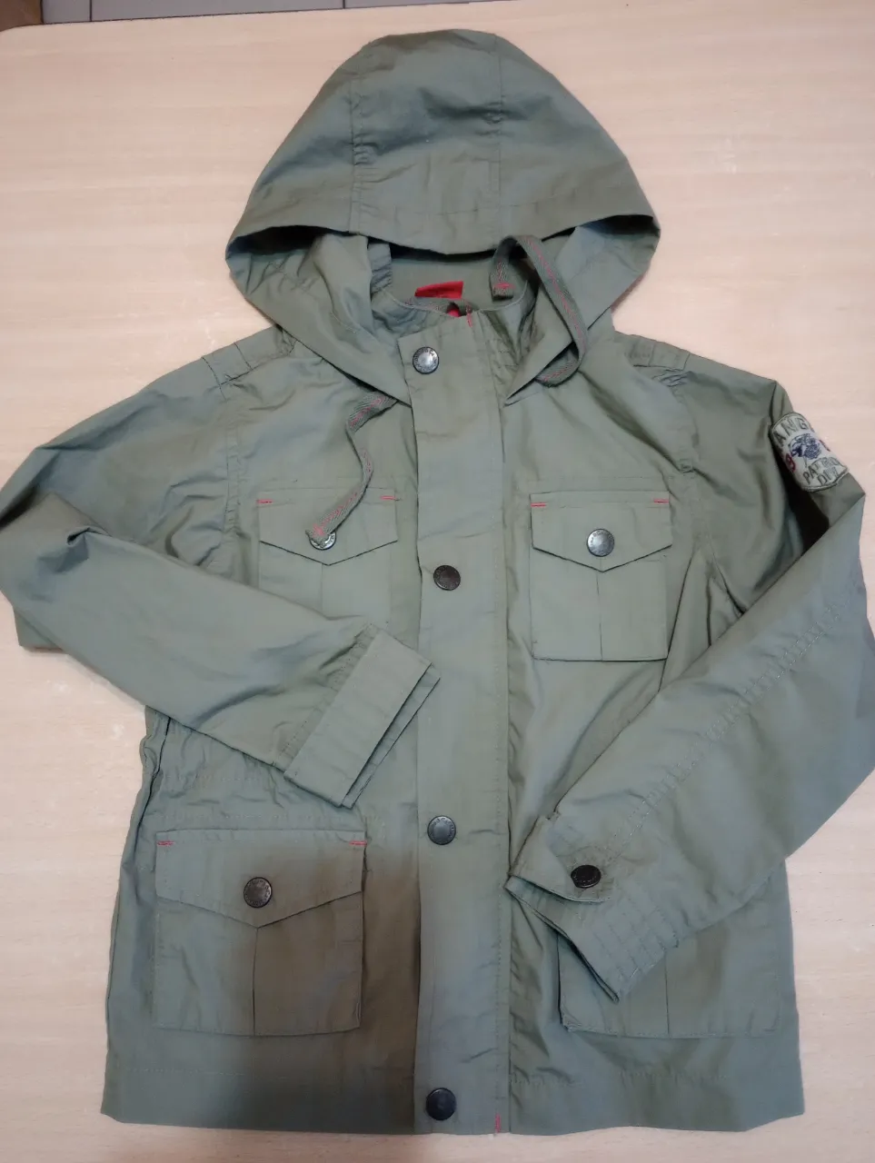 Campera verde con capucha para niño, estilo utilitario. Cuenta con cierre, bolsillos y detalles en rojo. Ideal para un look casual y cómodo.