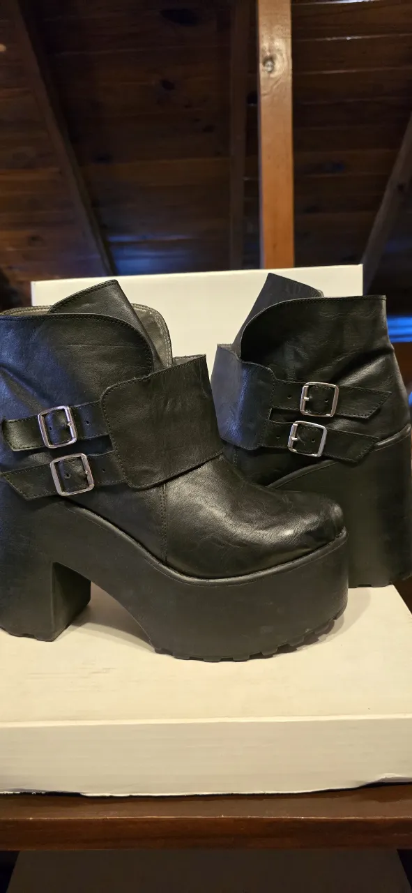 Botas tipo borcegos,  negras de cuero con plataforma alta y taco grueso. Tienen dos hebillas plateadas en la parte superior y una correa con hebilla en el lateral, zuela tractor 