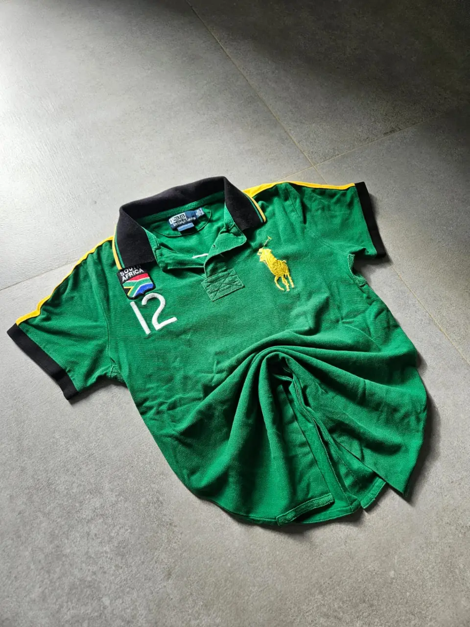 Remera polo de hombre de la marca Polo by Ralph Lauren, de color verde con detalles amarillos y negros. Tiene el número 12 en blanco y el logo del equipo de rugby de Sudáfrica. Ideal para un look deportivo y casual.