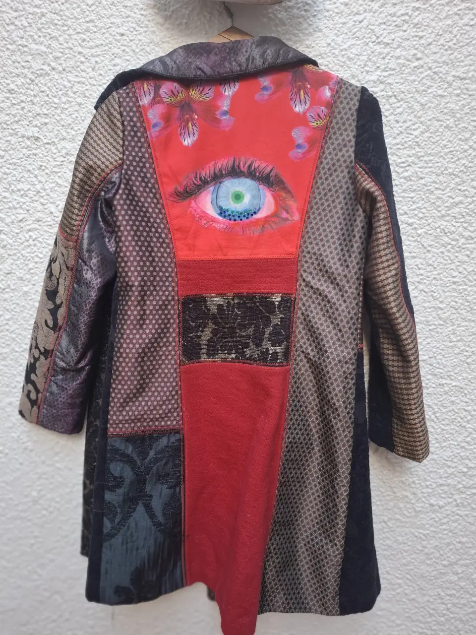 Tapado Desigual con diseño patchwork y detalles de ojo en la espalda. Forro interior rojo con estampado. Botones decorativos.