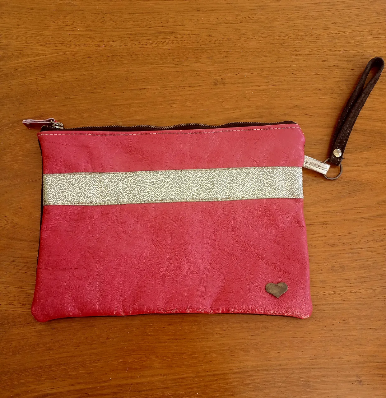 Cartera fucsia con una franja horizontal dorada texturizada en el centro. Posee un pequeño aplique metálico en forma de corazón en la parte inferior derecha y un detalle de cierre con tirador. Es de cuero vacuno. 