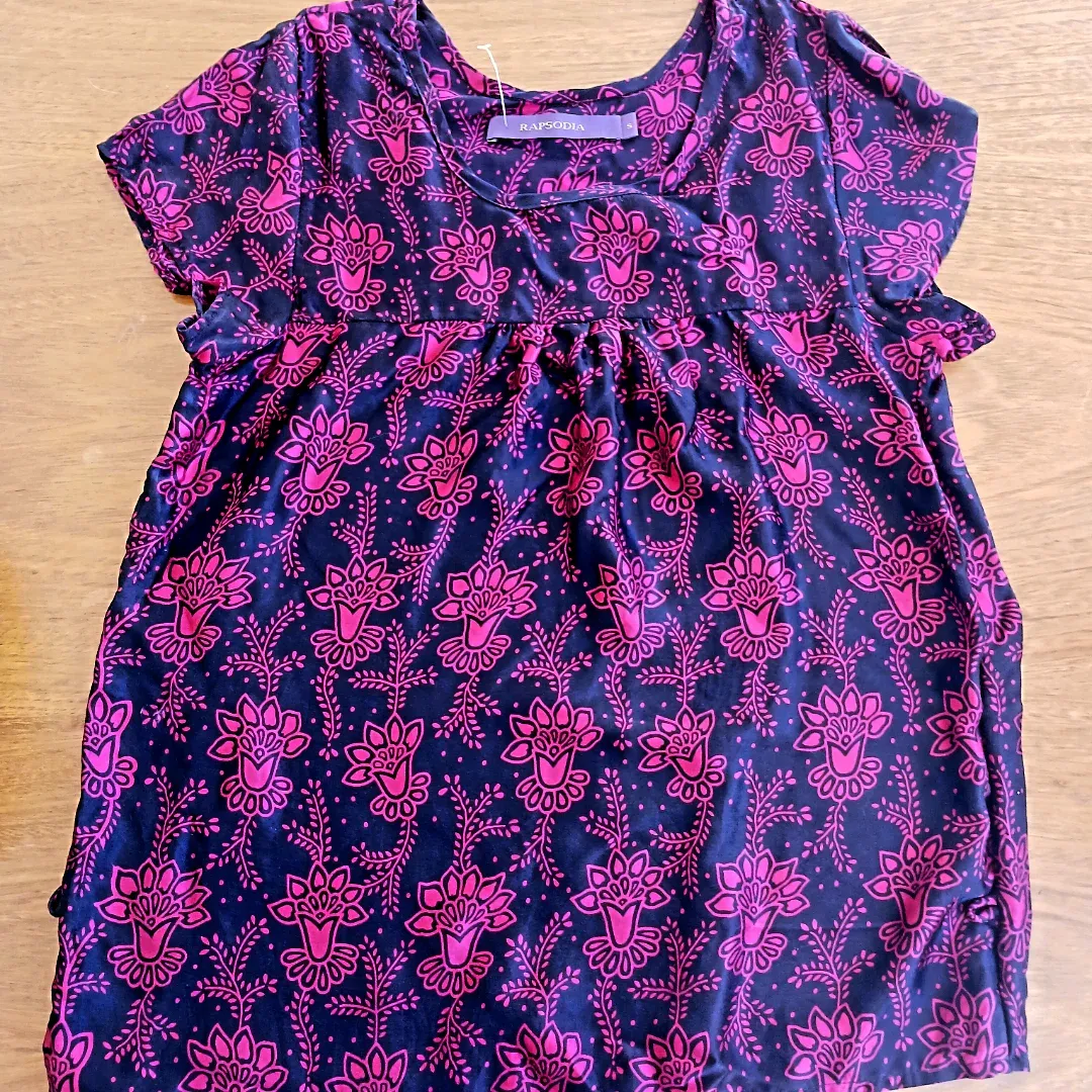 Blusa estampada con flores y hojas en color magenta sobre fondo negro. Tiene cuello redondo y mangas cortas.