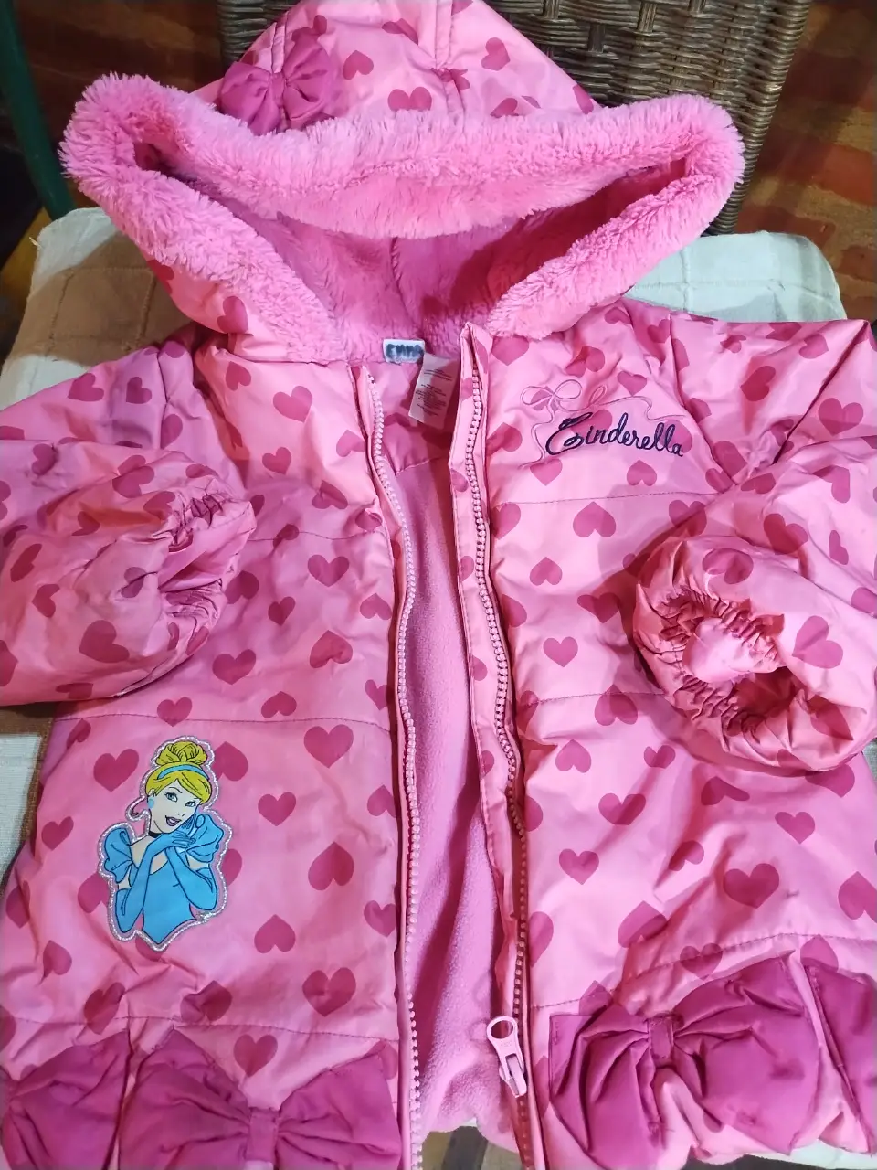 Campera rosa de niña con estampado de corazones y diseño de la princesa Cinderella. Cuenta con capucha con borde de piel sintética y detalles de lazo. Ideal para abrigar y lucir un estilo mágico.