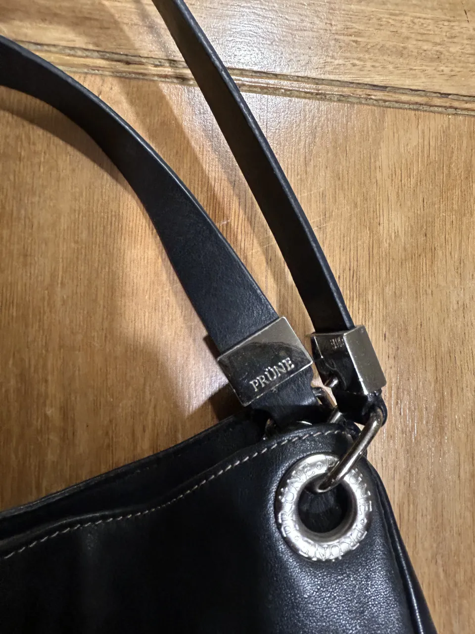 Bolso negro de cuero con correa de hombro. Tiene detalles metálicos en las uniones de la correa y un aro metálico en la parte superior.