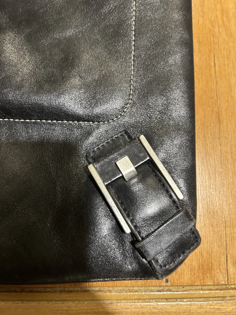 Cartera negra de cuero con correa larga y hebillas metálicas en la parte inferior. Posee un bolsillo frontal con cierre y otro bolsillo interno.