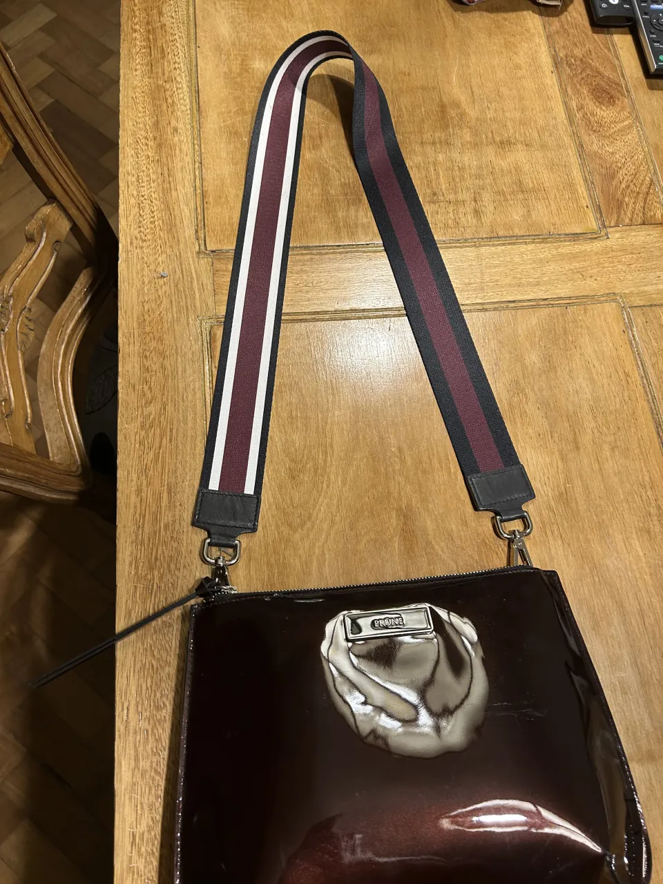 Bolso de cuero charolado color bordó con detalle de logo metálico en el frente. Correa de tela con franjas en color bordó, blanco y negro.