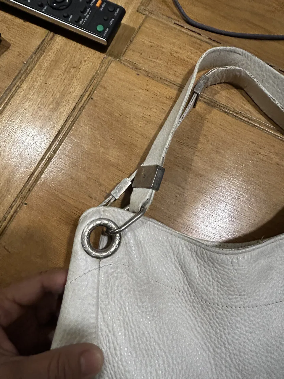 Bolso blanco de cuero con correa al hombro y placa metálica con el nombre de la marca.
