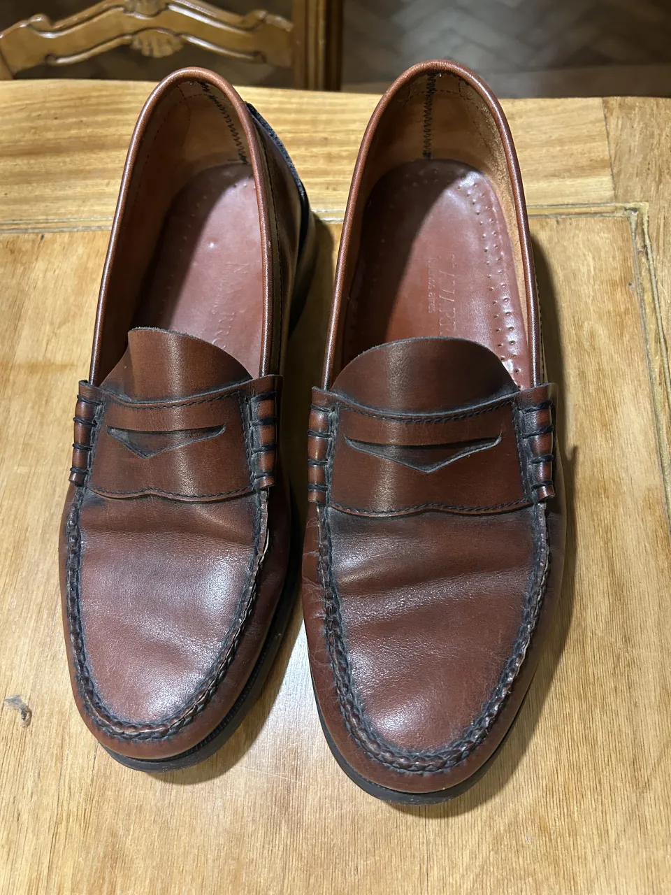 Zapatos mocasines de cuero color marrón. Tienen costuras visibles y un detalle de tira en la parte superior.