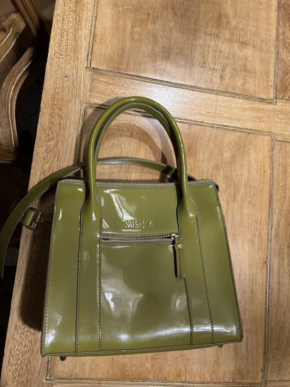 Bolso de mano color verde con correa larga desmontable. Tiene un bolsillo exterior con cierre y el logo de la marca en el frente. El interior cuenta con compartimentos.