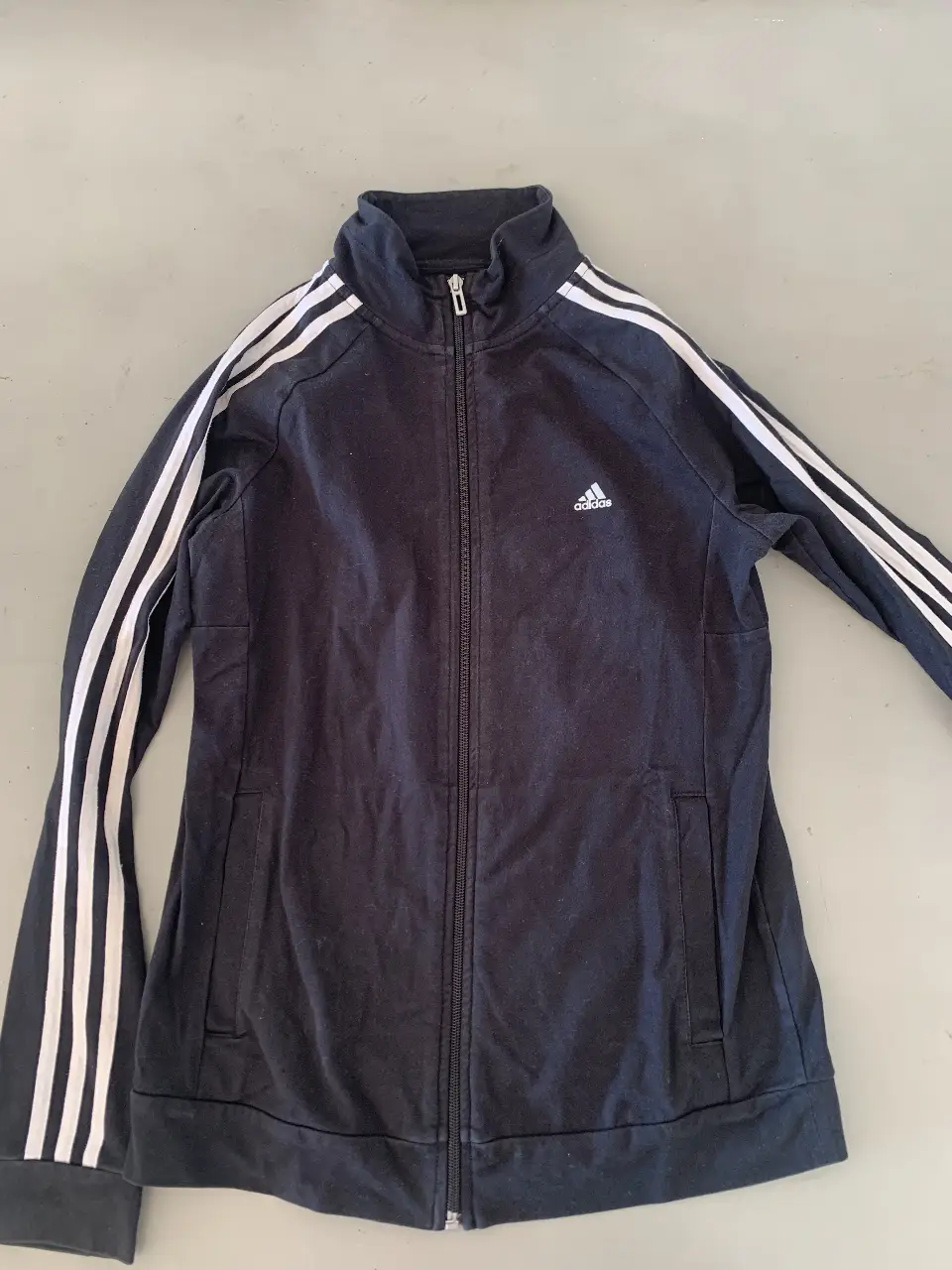 Buzo deportivo Adidas negro con cierre y las clásicas tres tiras blancas en las mangas. Ideal para un look casual y cómodo.