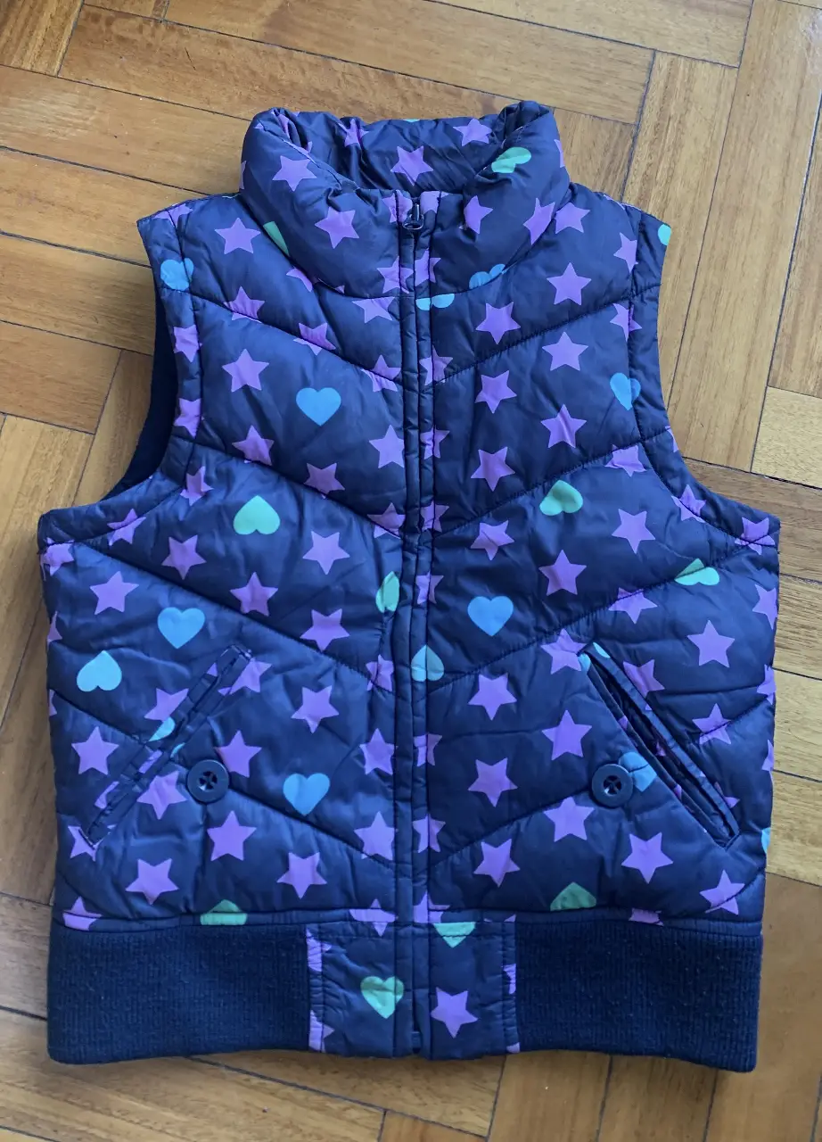 Chaleco acolchado para niña, con estampado de estrellas y corazones en tonos violeta, verde y celeste sobre fondo azul oscuro. Posee cierre frontal y bolsillos con botones.