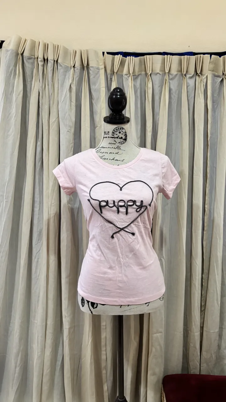 Remera manga corta de algodón color rosa pastel con estampa de corazón y la palabra "puppy" bordada en hilo negro. Ideal para un look casual y tierno.
