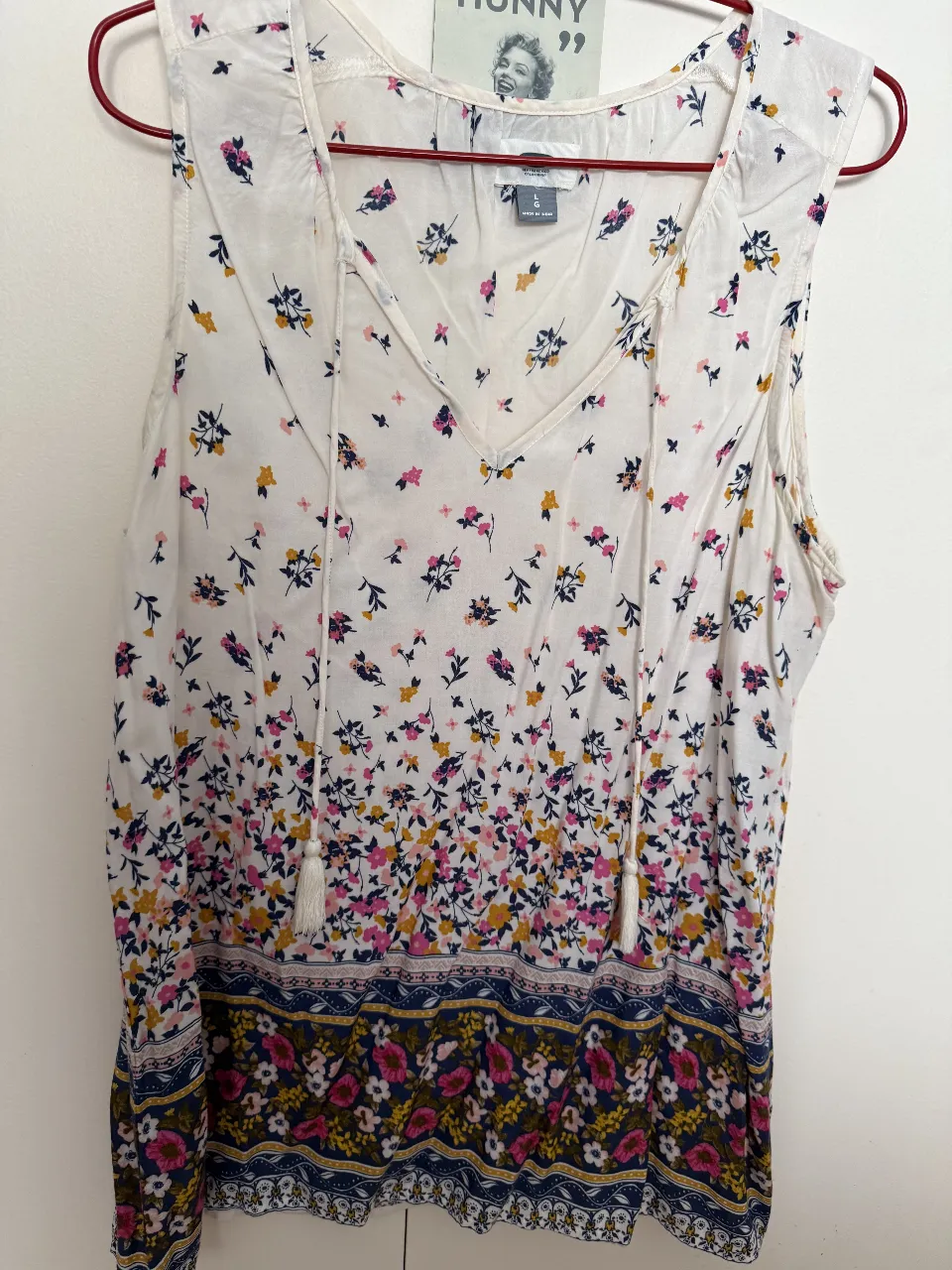 Musculosa blanca con estampa floral multicolor y detalle de guarda en tonos mas oscuros en la parte inferior. Posee tiritas con borlas en el escote en V.