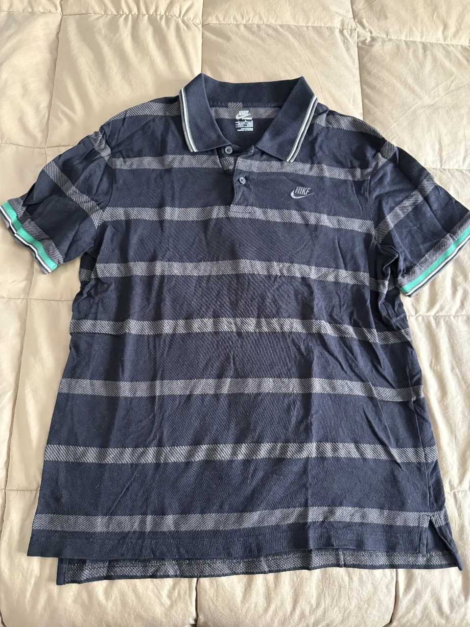 Remera polo de hombre, manga corta, con rayas horizontales en tonos de azul y gris. Detalle de cuello y puños con vivos en verde. Logo de la marca bordado en el pecho.