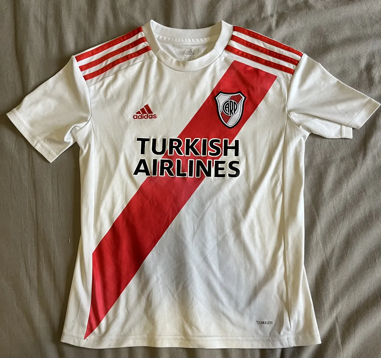 Camiseta de fútbol del River Plate, con el diseño tradicional de la banda roja en el pecho. Cuenta con el logo de Adidas y el patrocinador Turkish Airlines. Ideal para fanáticos y coleccionistas.