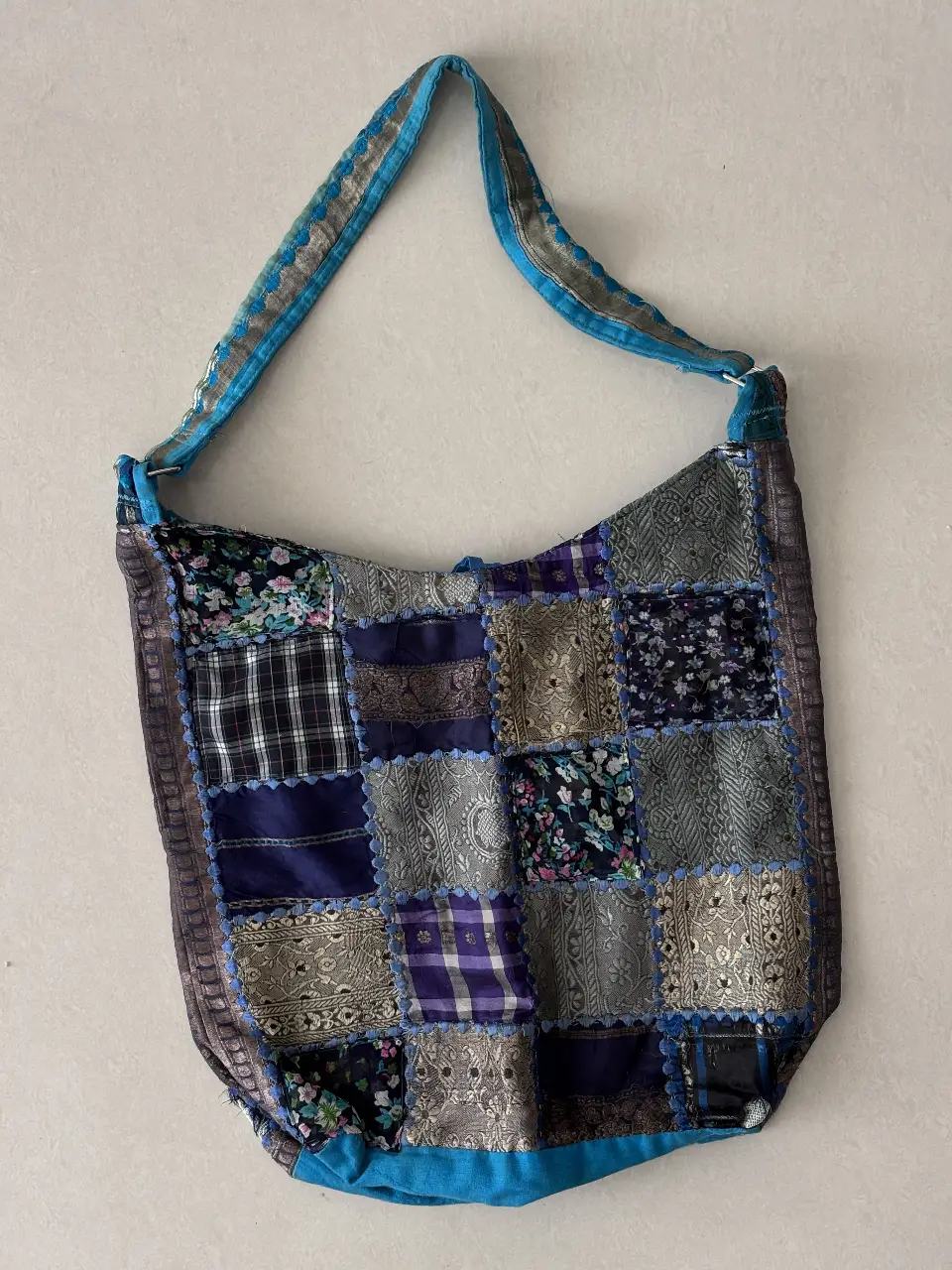 Bolso artesanal tipo bandolera, confeccionado con retazos de tela patchwork en tonos azules, grises y plateados. Detalles de bordado y cinta en el borde y la correa. Ideal para un look bohemio y original.