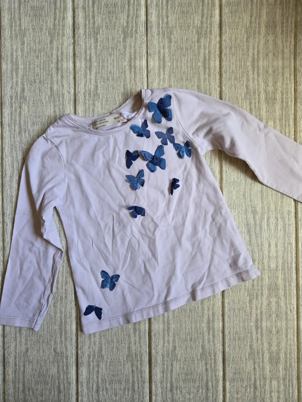 Remera manga larga de algodón color blanco con apliques de mariposas azules en relieve en el frente. Cuello redondo.