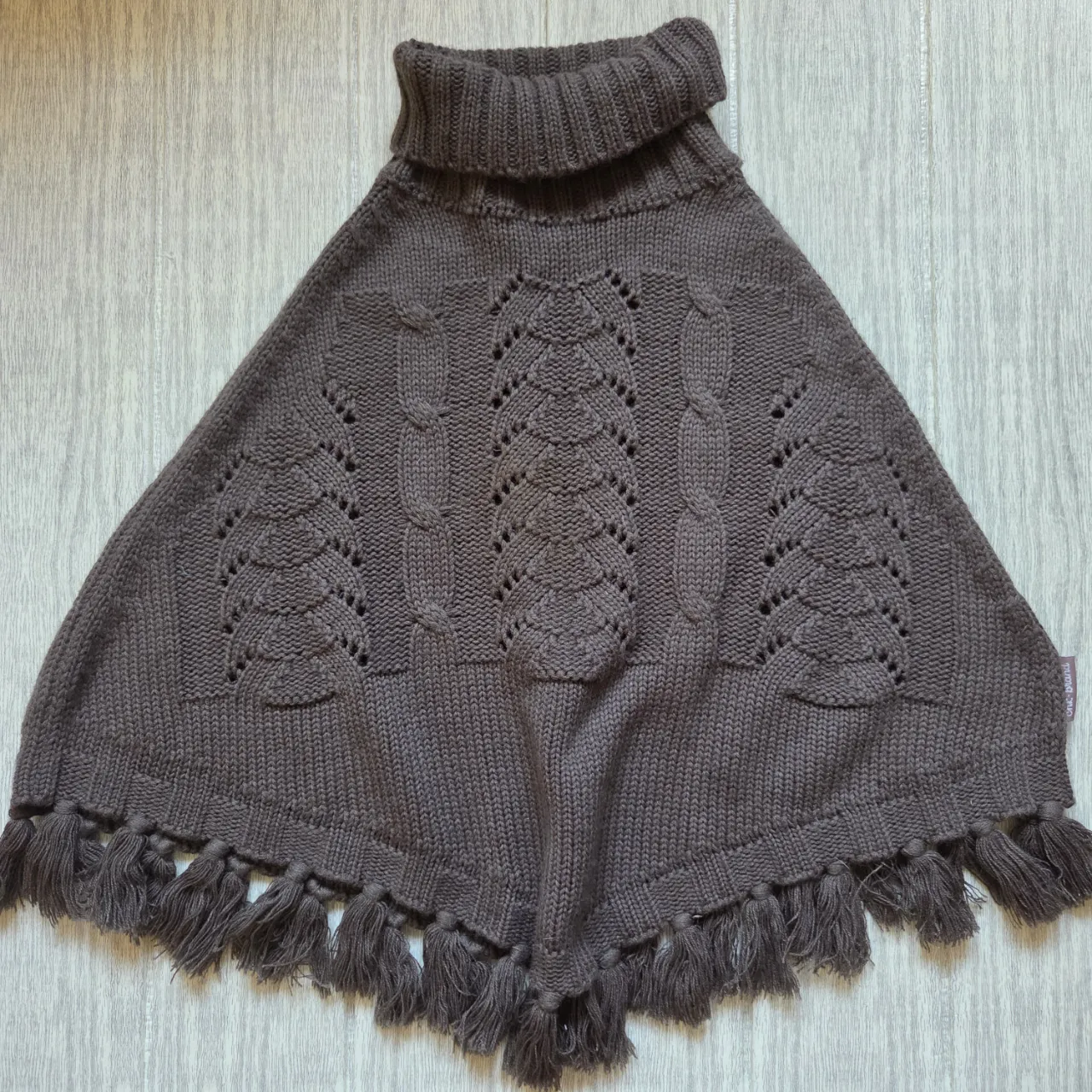 Poncho de tejido marrón con cuello alto y detalles de trenzas y hojas caladas. Posee flecos en la parte inferior.