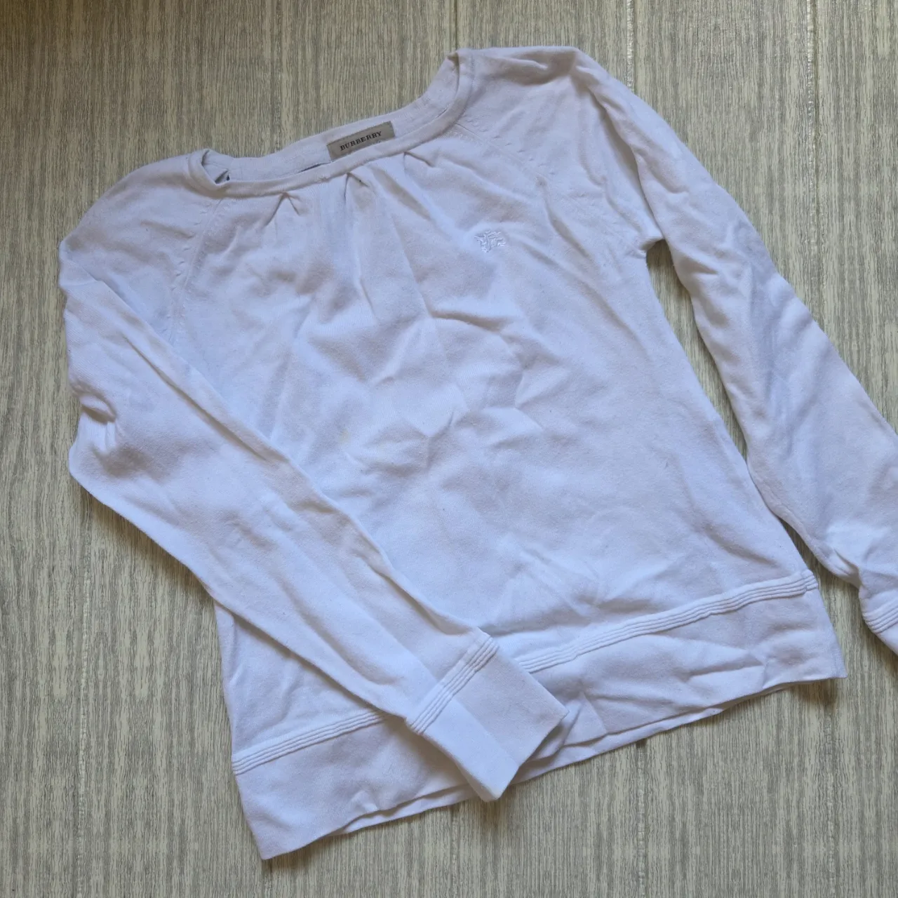 Remera blanca de manga larga con cuello redondo y detalle fruncido en la parte superior. Presenta un pequeño logo bordado en el frente.