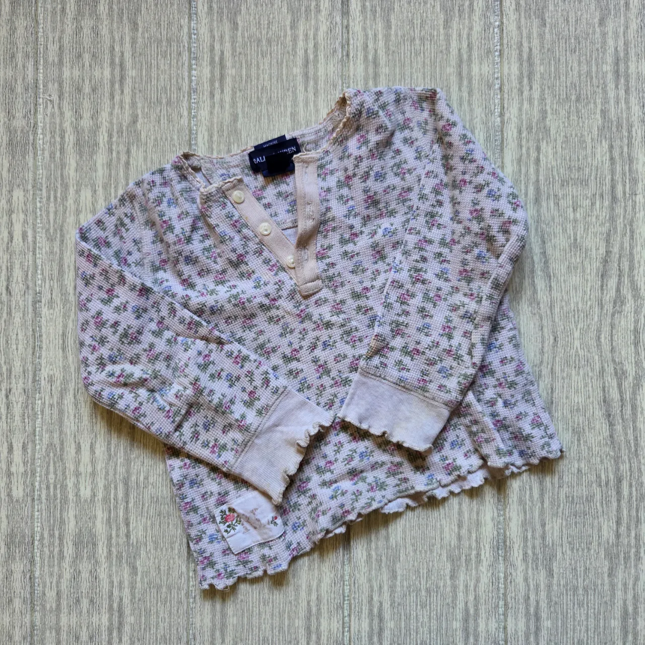 Remera manga larga de algodón con estampa floral de flores rosas, azules y verdes sobre fondo blanco. Posee cuello redondo con botones y puños y ruedo con terminación de ondas.