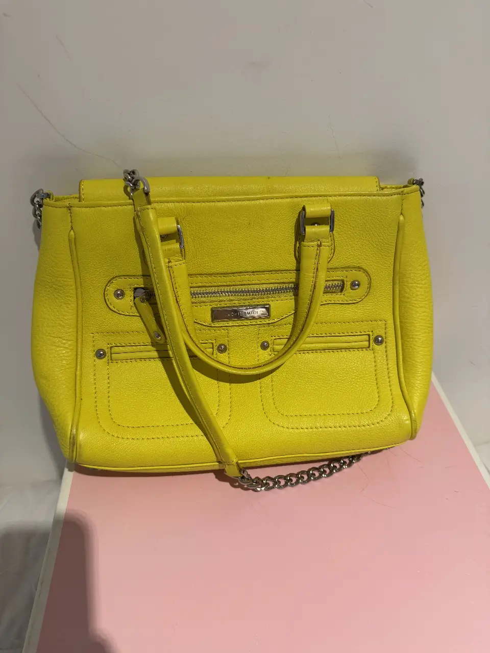 Cartera amarilla de cuero, con manijas y correa para colgar. Tiene detalles plateados y un bolsillo frontal con cierre. Ideal para un look casual y moderno.