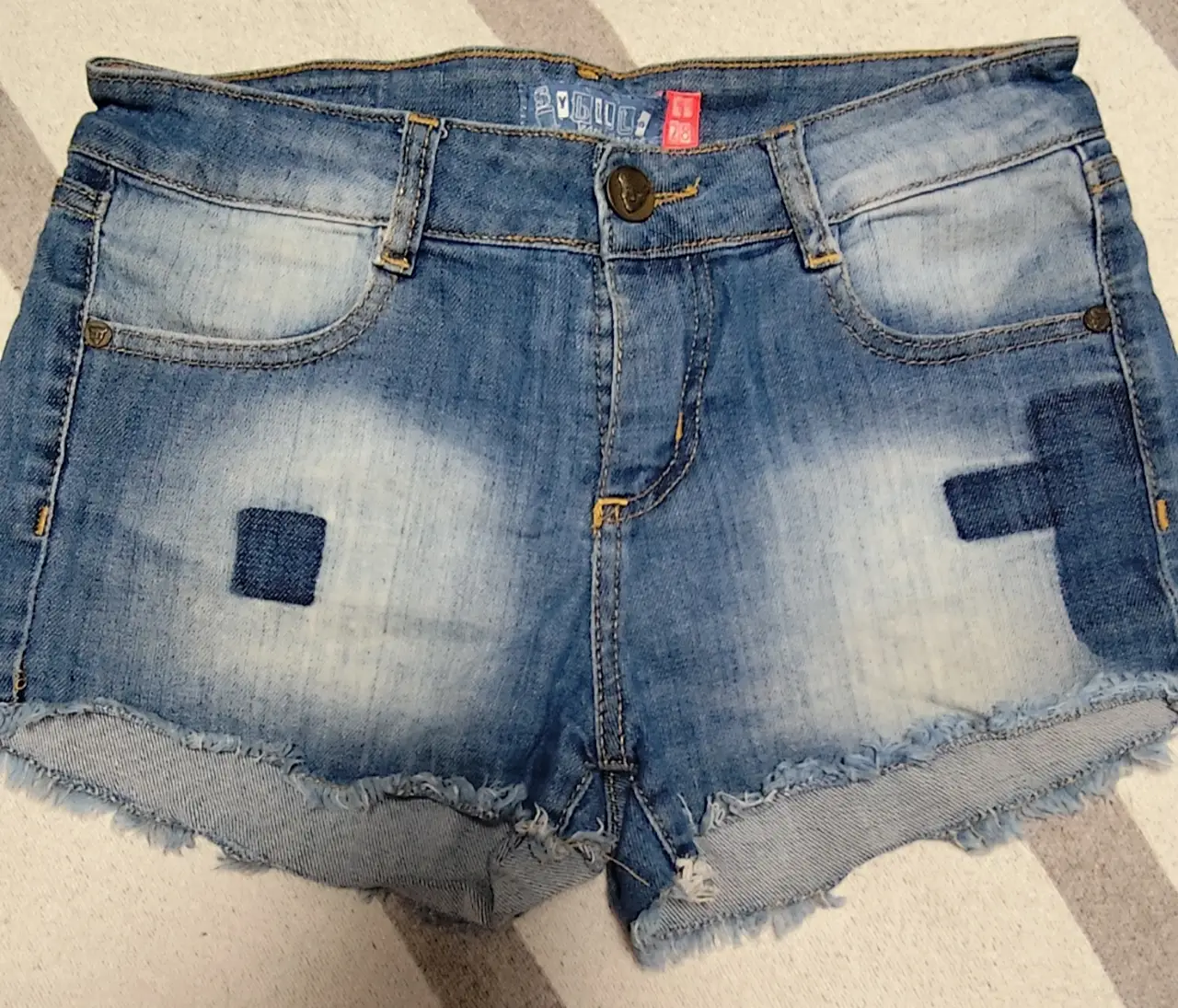 Short de jean de mujer, tiro medio, con detalles de parches en la parte delantera. Ideal para un look casual y moderno.