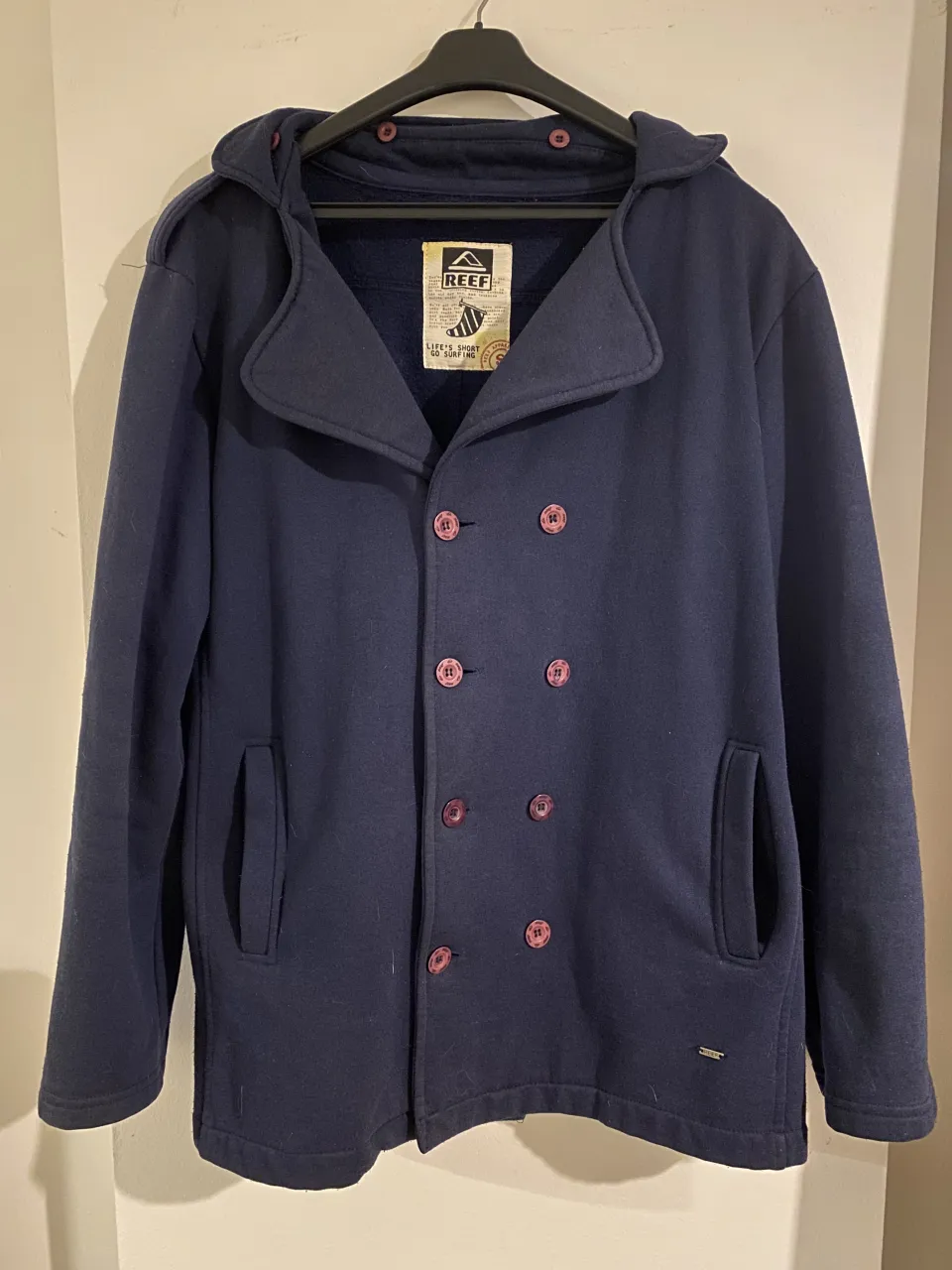 Campera azul marino con capucha, doble abotonadura y cuello solapa. Posee dos bolsillos frontales y botones color rosa.