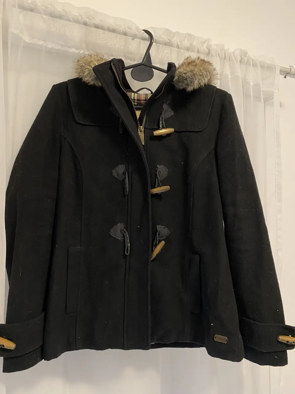 Tapado negro con capucha desmontable y detalles de madera. Ideal para el invierno, con un estilo clásico y abrigado. Cuenta con un forro interior a cuadros. con detalles de uso como se ve en la foto 