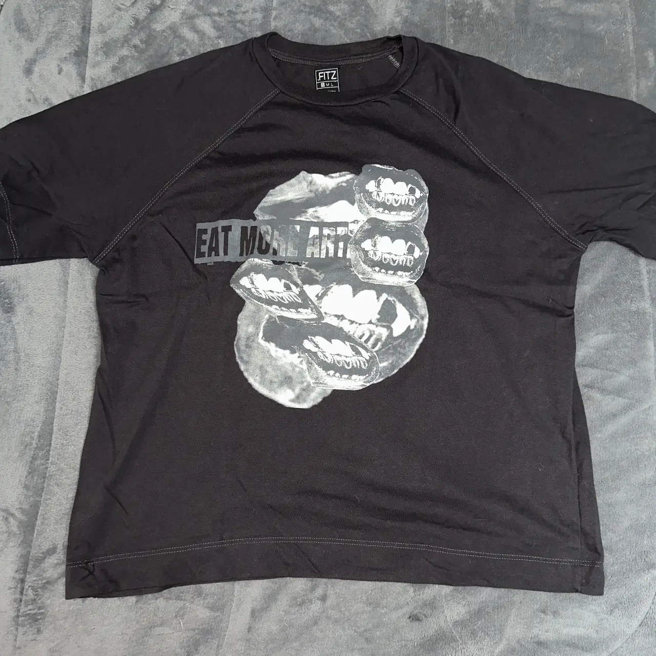 Remera gris de manga corta con estampado "EAT MORE ART" en gris. Diseño con bocas y dientes. Ideal para un look urbano y moderno.