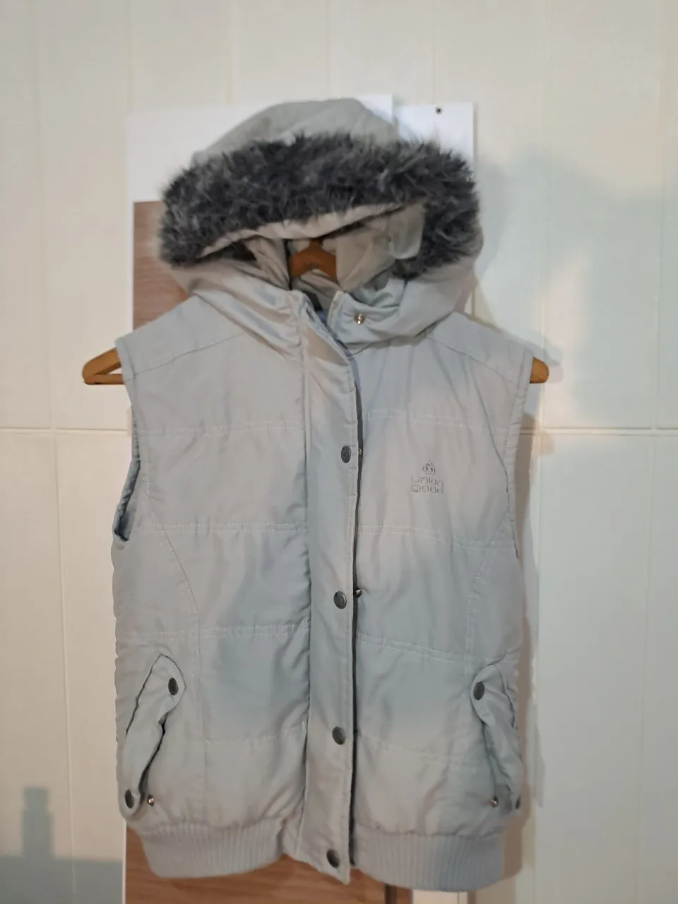 Chaleco acolchado gris con capucha forrada en piel sintética. Cierre de broches frontales y bolsillos laterales con solapa.