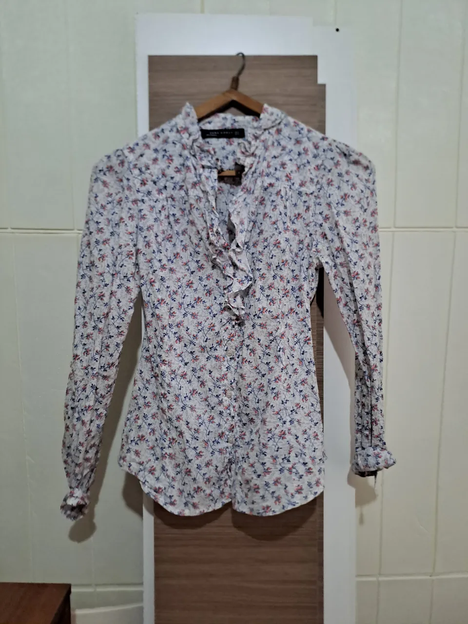 Camisa de zara estampada con flores floreada pequeñas en tonos rojos y azules sobre fondo blanco. Posee cuello con volados y detalles de volados en la parte delantera central. Mangas largas con puños abotonados.