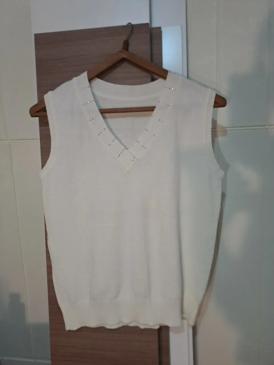 Sweater sin mangas de tejido blanco con escote en V. El borde del escote tiene detalles de brillitos plateados.