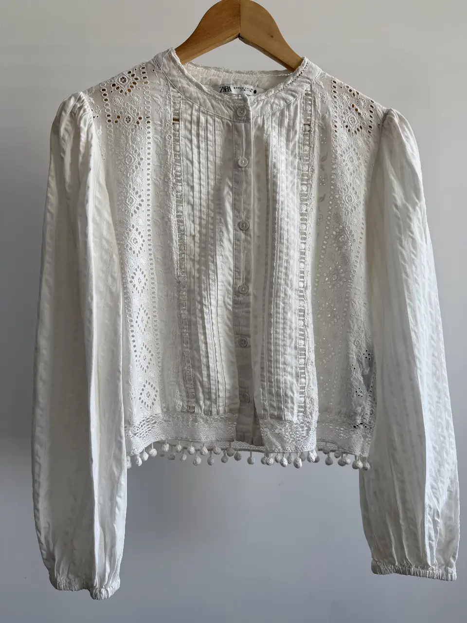 Blusa blanca de encaje con detalles de pompones en el borde inferior. Diseño elegante y romántico, ideal para un look casual o semi-formal.