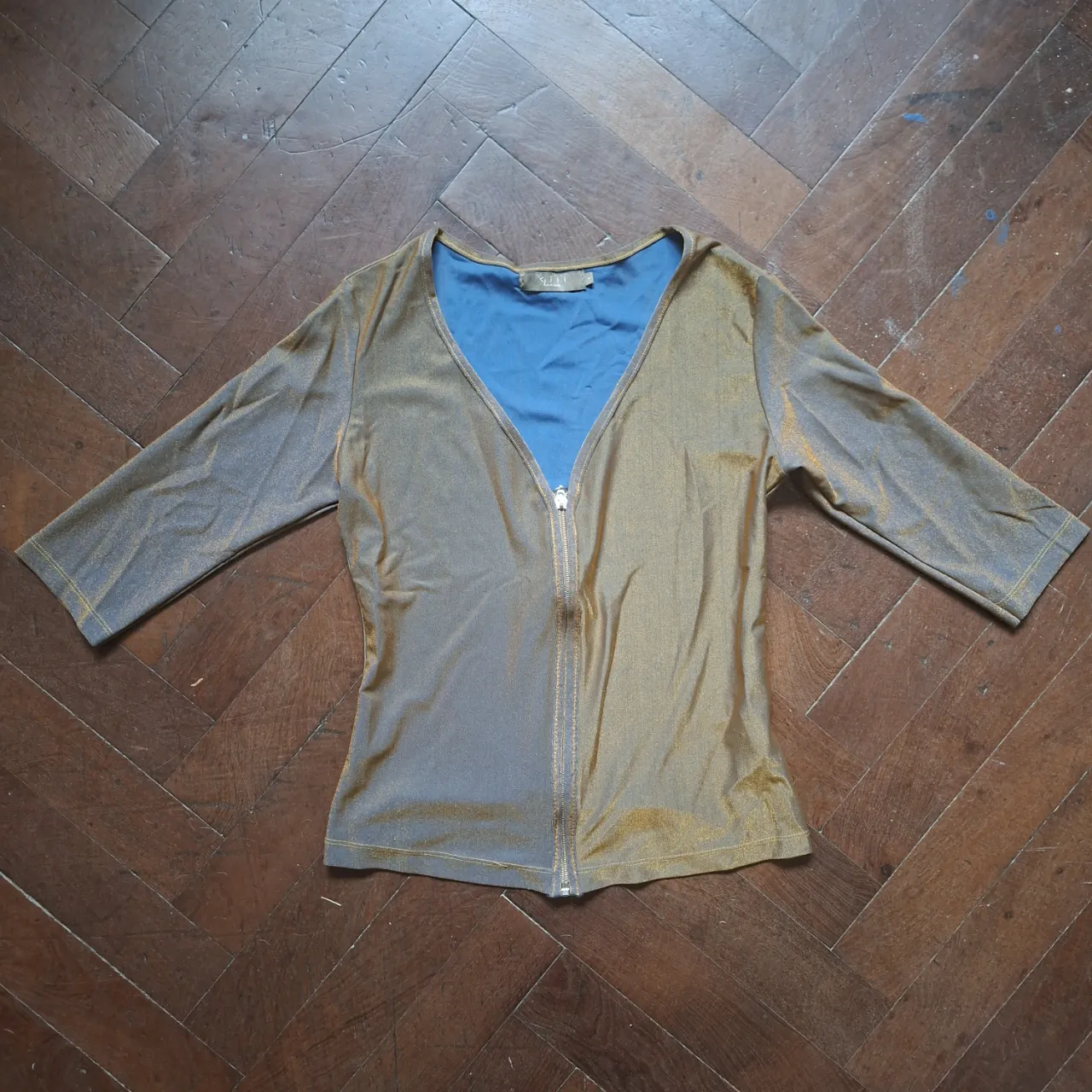 Remera manga 3/4 con cierre al frente y cuello en V. Posee un tejido brillante con detalles en dorado y azul.
