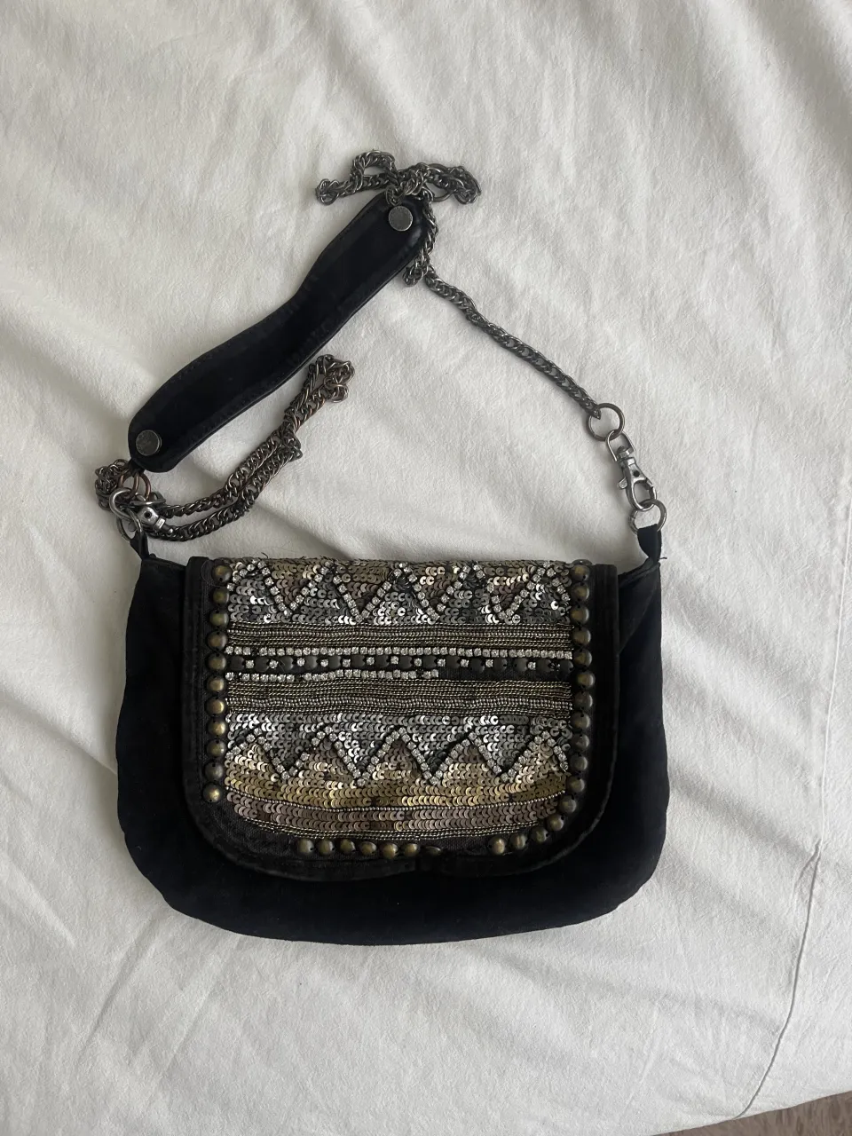 Cartera negra con detalles de lentejuelas y tachas plateadas y doradas en la solapa. Correa de cadena metálica.
