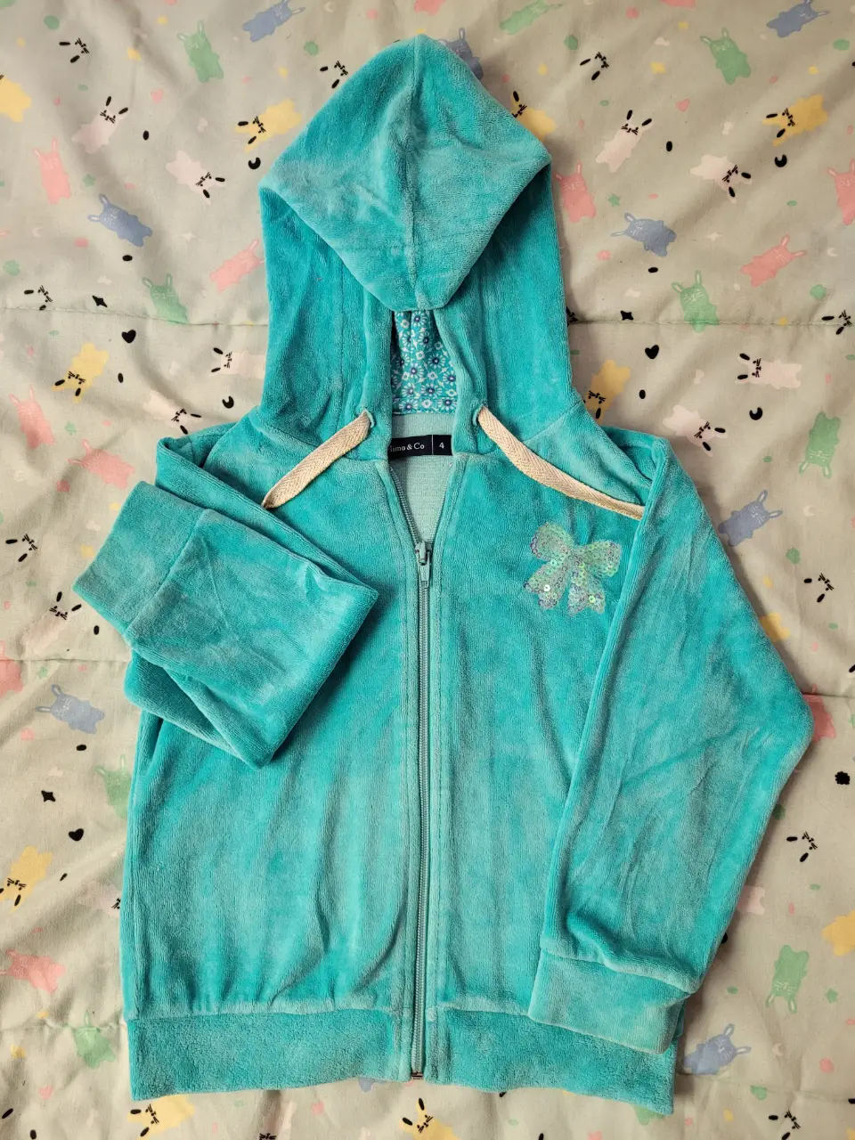 Buzo con cierre de niña, color celeste, con capucha y detalle de moño con lentejuelas. Ideal para un look casual y abrigado.
